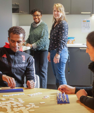 twee mensen spelen een spel aan tafel, twee mensen kijken blij vanuit de keuken
